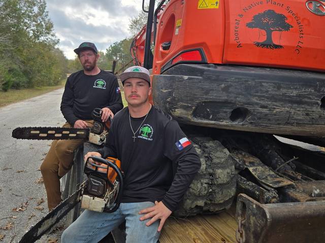 Two crew members holding chainsaws, seated on the tracks of an excavator marked with the company logo