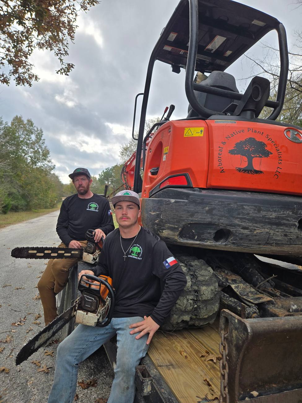 Two crew members holding chainsaws, seated on the tracks of an excavator marked with the company logo