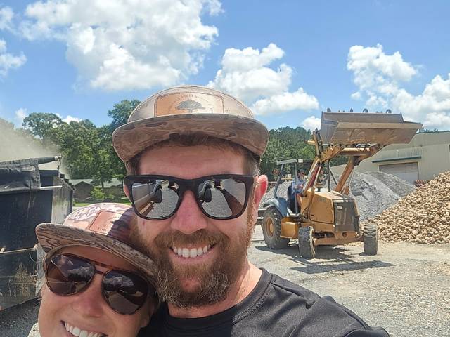 Two crew members at a materials yard, with a loader working piles of crushed rock and gravel