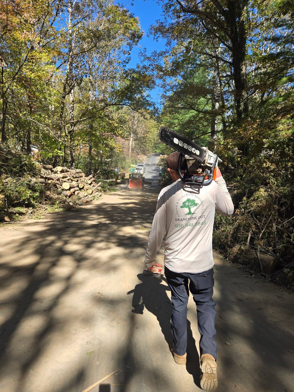 A crew member walking a road carrying a chainsaw, with cut storm debris stacked along the verge